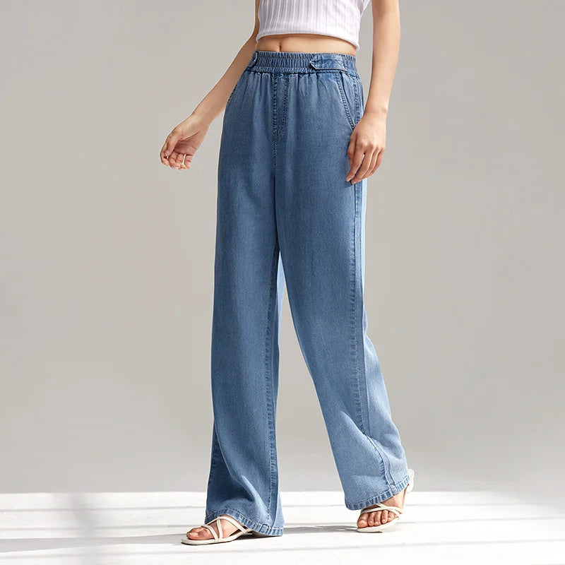 A person wearing light blue, wide-leg, breathable denim pants with an elastic waistband, a white sleeveless crop top, and white sandals stands against a plain, light background. Only the lower half of their body is visible.