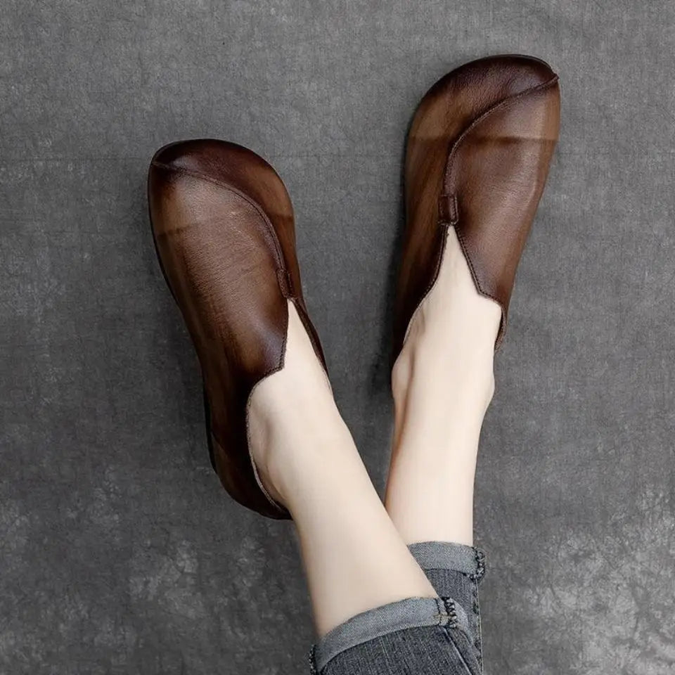 A person wearing brown slip-on shoes made from supple leather with a unique curved design, resting their feet on a gray textured surface. The person is dressed in rolled-up, faded blue jeans.