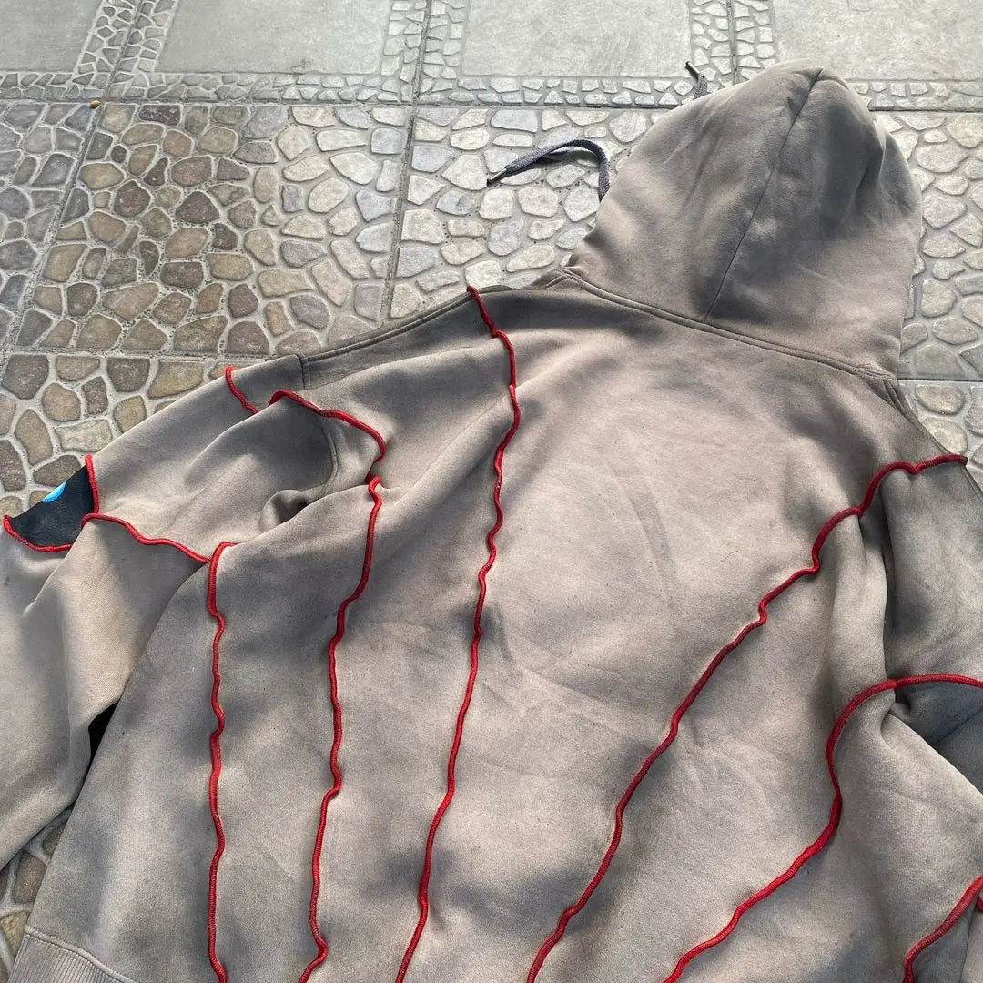 A beige hoodie with a hood and red wavy stitched lines along the back and sleeves, crafted from breathable fabric, is laid flat on a tiled surface with a stone and geometric pattern.