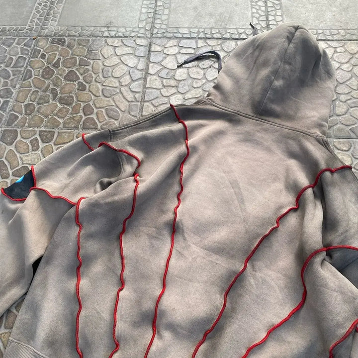 A beige hoodie with a hood and red wavy stitched lines along the back and sleeves, crafted from breathable fabric, is laid flat on a tiled surface with a stone and geometric pattern.