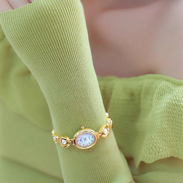 A person wearing a light green long-sleeved top displays a gold wristwatch with pearl accents. The watch features an oval face with a white dial, precision quartz movement, and is stylishly worn over the sleeve.