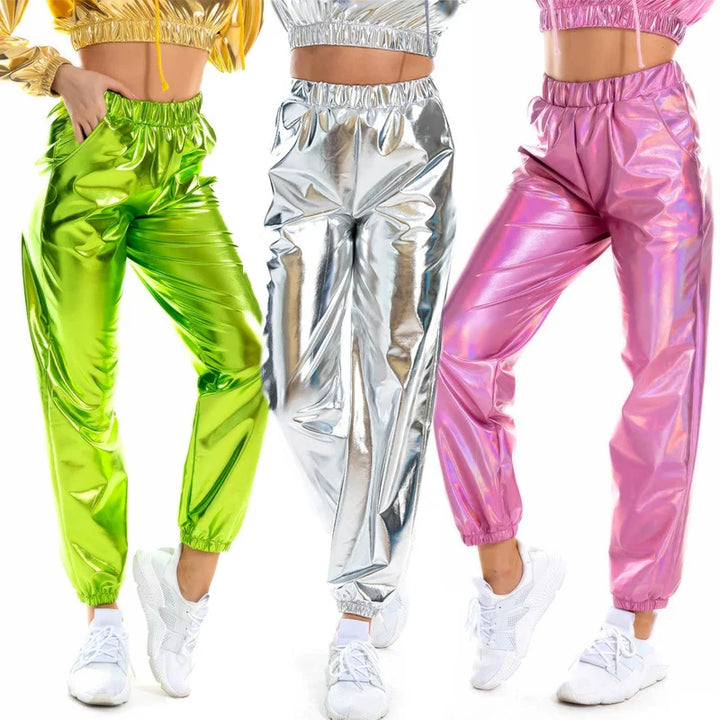 Three people wear shiny jogger pants with an elastic high waist and loose fit in metallic green, silver, and pink, each with matching cropped jackets. All sport white sneakers and pose against a white background.