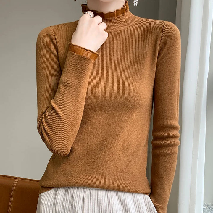 A person wearing a fitted brown long-sleeve top with a ruffled mock neck and cuffs, paired with a light pleated skirt, standing indoors near a window.