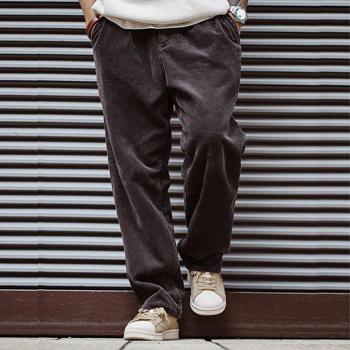 A person stands against a striped metal wall, hands in pockets, wearing loose dark brown corduroy pants, a cream sweater, a wristwatch, and beige Lowland Odyssey Sneakers—their head is not visible in the image.