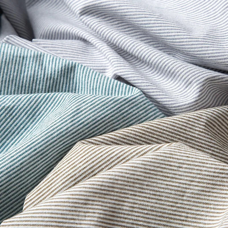 Close-up of several pieces of fabric with diagonal pinstripes in light blue, gray, beige, and off-white, reminiscent of a classic pinstripe shirt, draped and overlapping each other.