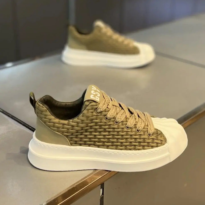 A stylish olive-green woven upper sneaker with a cushioned sole and matching laces is displayed on a reflective surface; another similar shoe is blurred in the background.