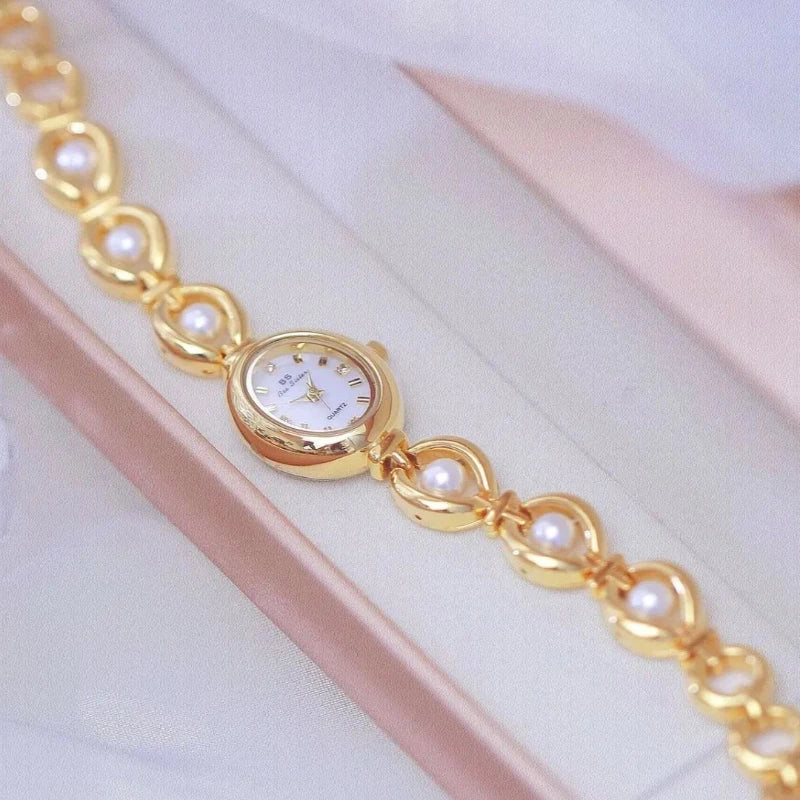 A gold wristwatch with a white round face and pearl accents is displayed on a soft pink and white background. Featuring precision quartz movement, the bracelet’s alternating gold links and pearls give it an elegant, jewelry-like appearance.