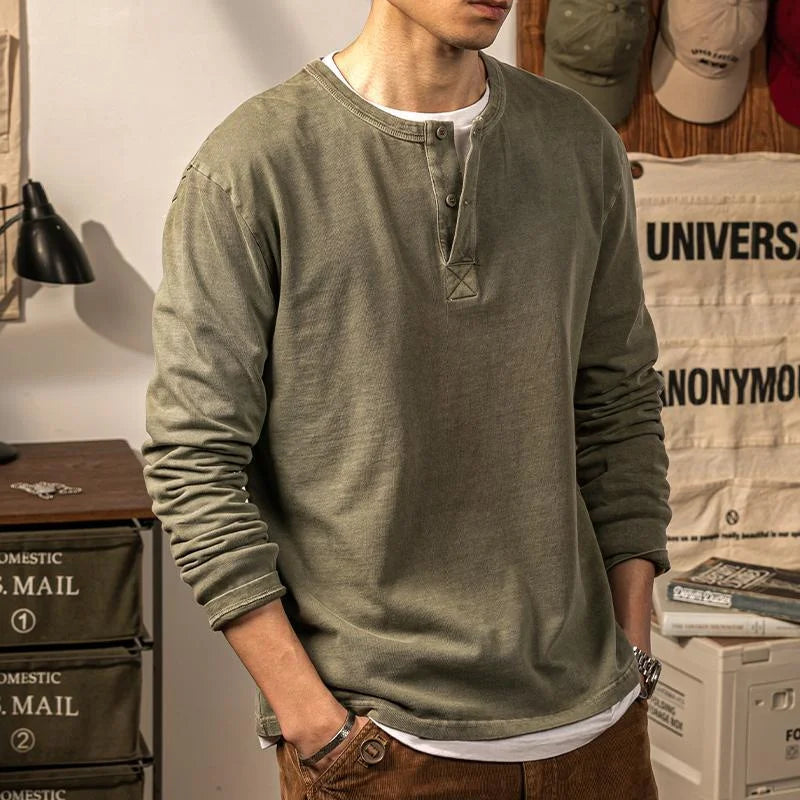 A person wearing a pure cotton olive green long-sleeve Henley shirt with brown pants stands indoors, hands in pockets. The background includes storage drawers, hats, and hanging canvas with printed text. The persons face is not visible.