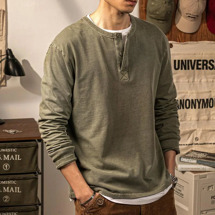 A person wearing a pure cotton olive green long-sleeve Henley shirt with brown pants stands indoors, hands in pockets. The background includes storage drawers, hats, and hanging canvas with printed text. The persons face is not visible.