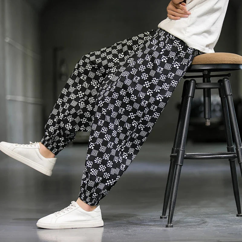 A person sits sideways on a stool wearing black cotton joggers with a white geometric pattern, a white shirt, and white sneakers—making a subtle fashion statement against a minimal, blurred background.