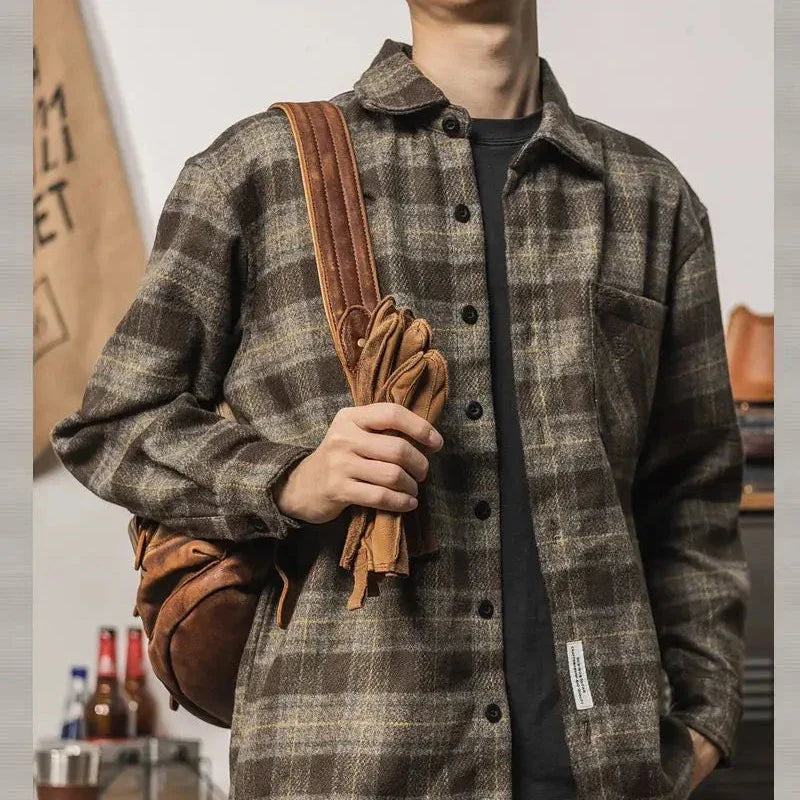 A person wearing a relaxed fit brown and gray plaid button-up shirt holds the strap of a brown leather backpack slung over their shoulder, with gloves attached. The persons face is not visible.