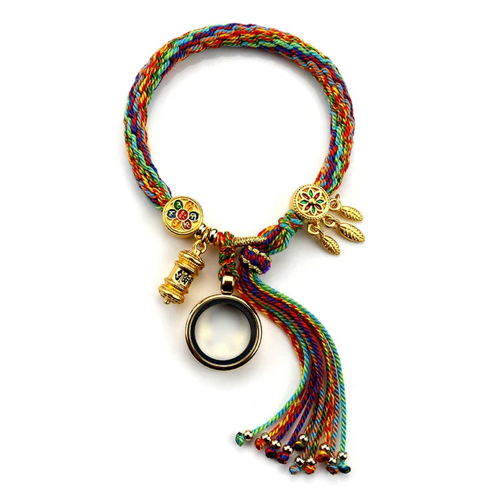 A colorful braided bracelet with gold charms, including a ring, a cylindrical charm, and a flower with dangling leaves. Showcasing Tibetan craftsmanship, the adjustable bracelet features multicolored threads and a tasseled end.
