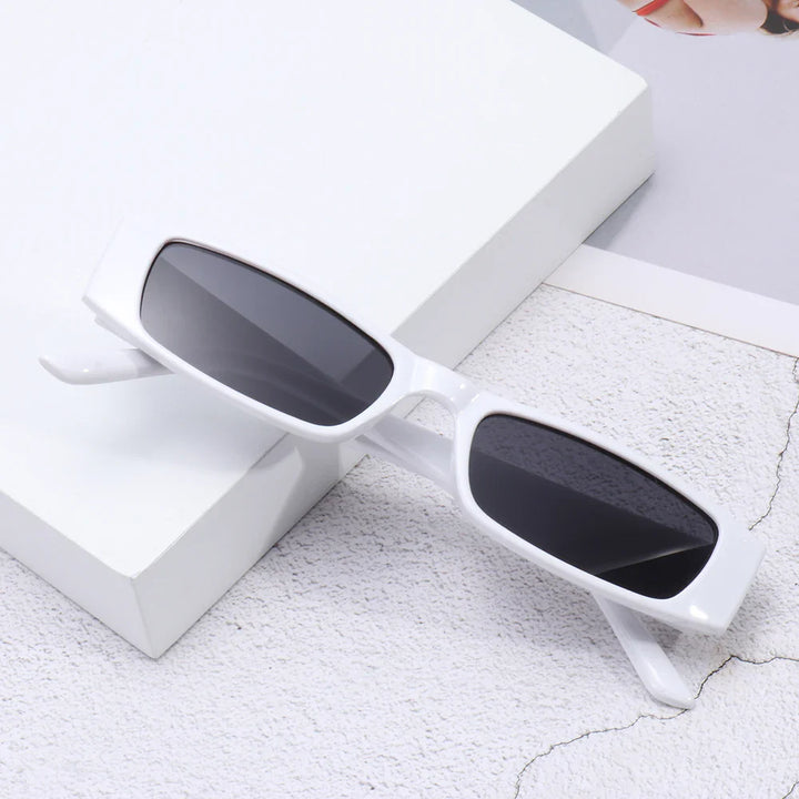 White retro rectangle sunglasses with dark lenses rest on a textured light surface, partially overlapping a white box and a corner of a magazine. The lightweight frame offers comfort and UV400 protection.