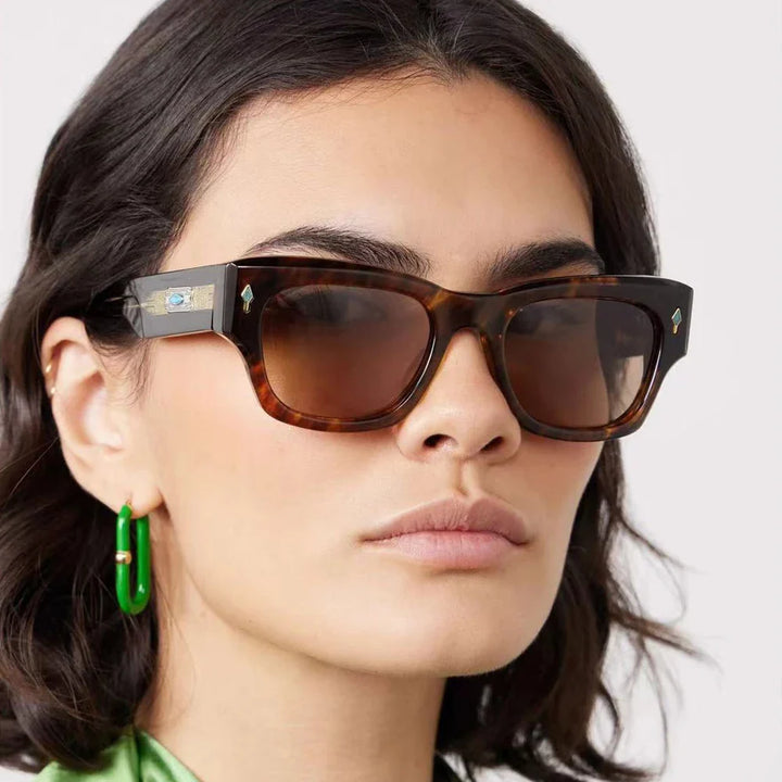 A woman with wavy dark hair wears vintage-inspired sunglasses with tinted lenses and green earrings. She has a neutral expression and is dressed in a shiny green top.