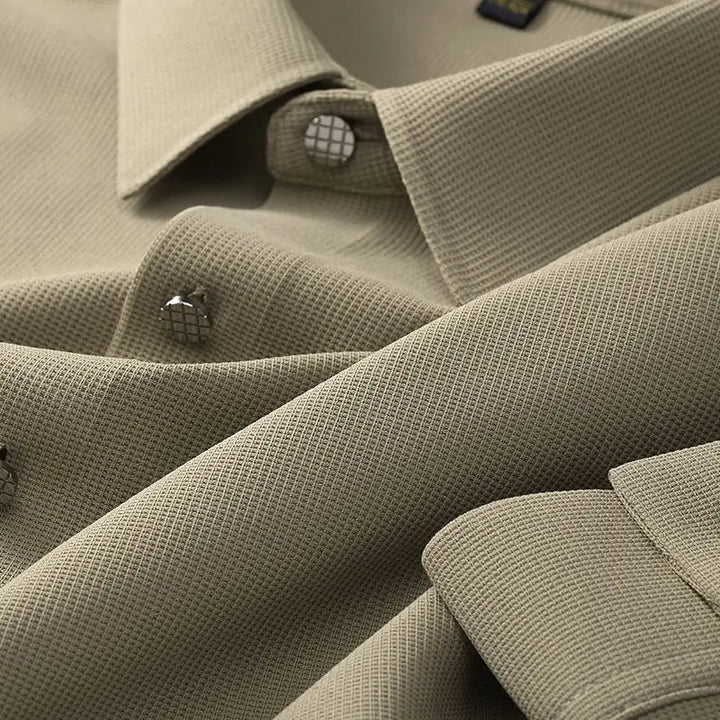 Close-up of a beige wrinkle-resistant shirt with a grid-like pattern, featuring metal buttons and visible collars and cuffs. The fabric appears smooth and finely woven.