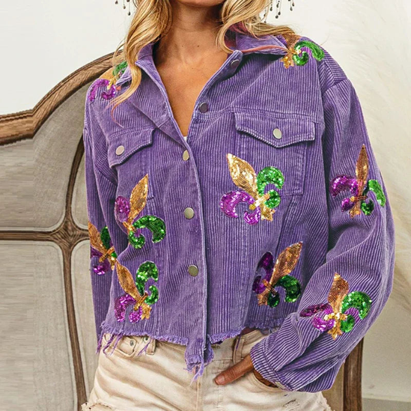 A woman wears a cropped, relaxed-fit purple corduroy jacket adorned with gold, green, and purple sequin floral patches, paired with light distressed shorts.