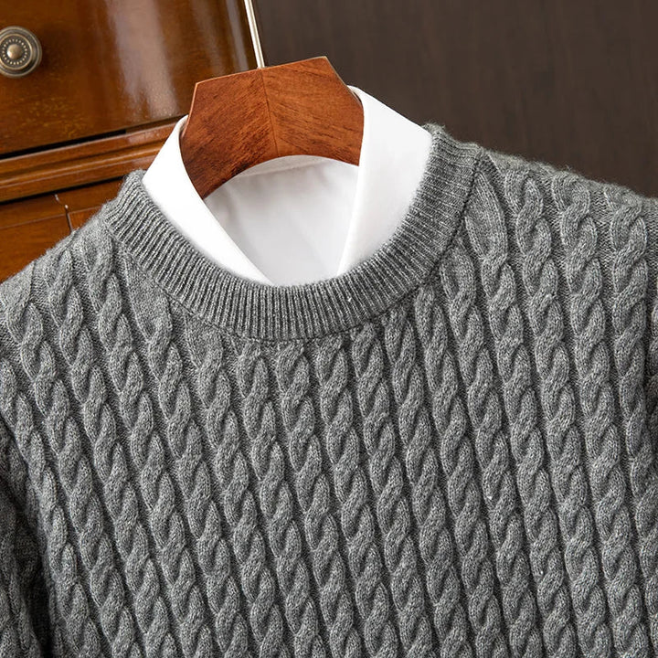 A close-up of a gray cable knit cashmere sweater with ribbed cuffs, layered over a white collared shirt, displayed on a wooden hanger against a dark brown background.