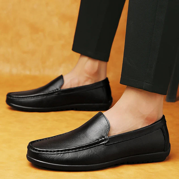 A person wearing black leather slip-on loafers with a cushioned footbed and black pants stands on a yellowish-brown surface, showcasing the shoes in a side view.