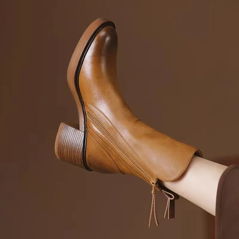 A close-up of a person wearing tan leather ankle boots with a chunky, low heel and side zipper, set against a plain brown background—perfect women’s boots for the season.