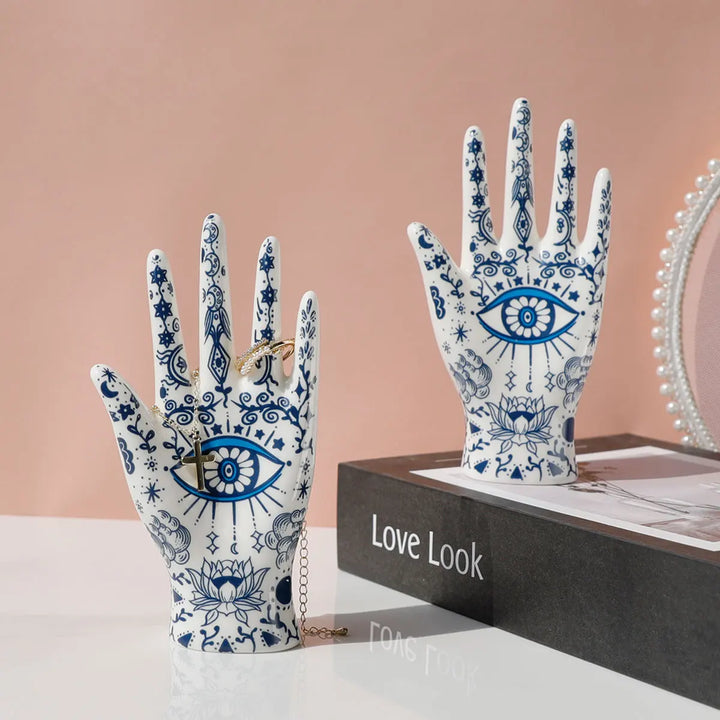 Two white ceramic jewelry stands with blue evil eye and floral designs hold jewelry, displayed on a book titled Love Look, with a round, pearl-framed mirror in the background—perfect for boho-chic home décor.