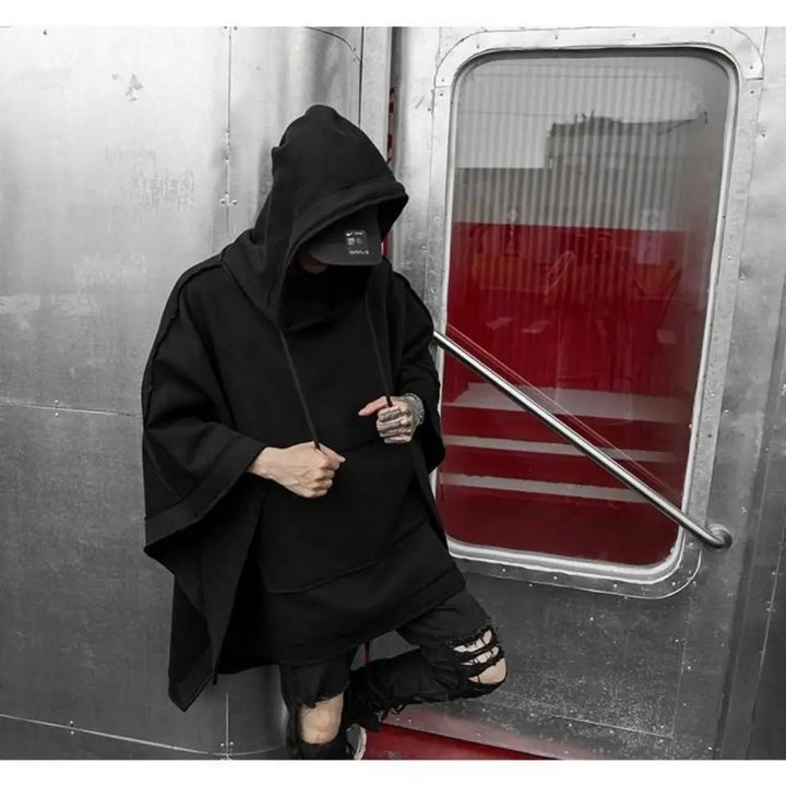 A person in a black Techwear hoodie and face mask stands against a metallic wall and red door, one hand holding the hoodie strings and the other in a pocket. The streetwear outfit is completed with ripped black pants.