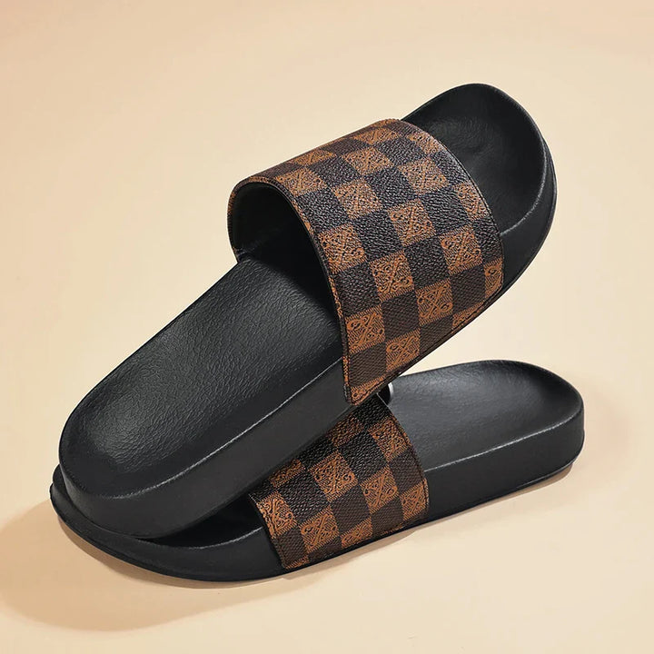 A pair of black streetwear slides with brown and black checkered straps, arranged in a stacked, crisscrossed position on a beige background.