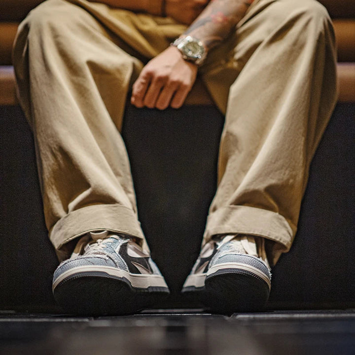 A person wearing beige pants and Denimwave 1986 Pro Sneakers sits with hands resting on their knees. The photo, focused on their shoes and lower legs, highlights the 80s aesthetic with softly blurred background.