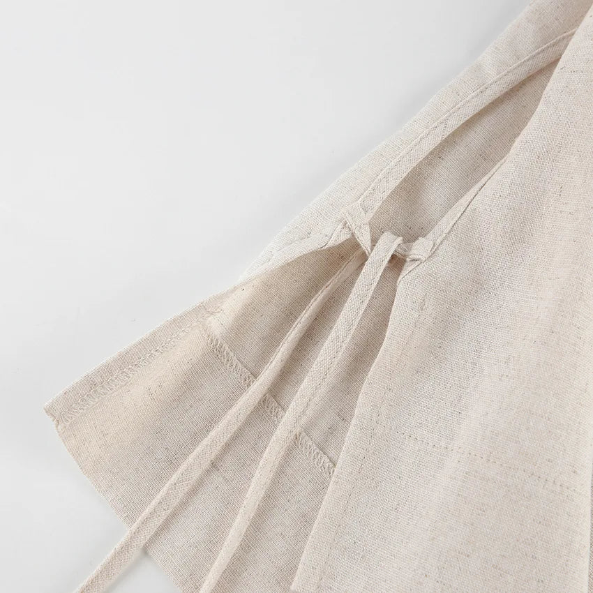Close-up of a beige Belgian Linen Blend apron with two fabric waist ties lying on a white surface. The texture of the material and seams are clearly visible.