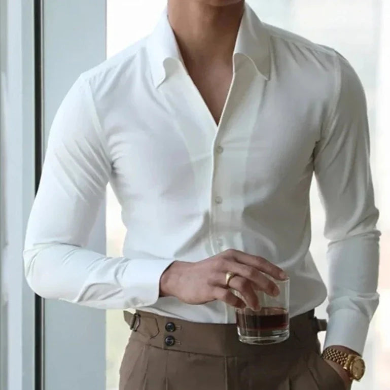 A man in a slim fit white dress shirt and brown trousers stands by a window, holding a glass of dark beverage. He wears a gold watch and a ring on his left hand. His face is not fully visible.