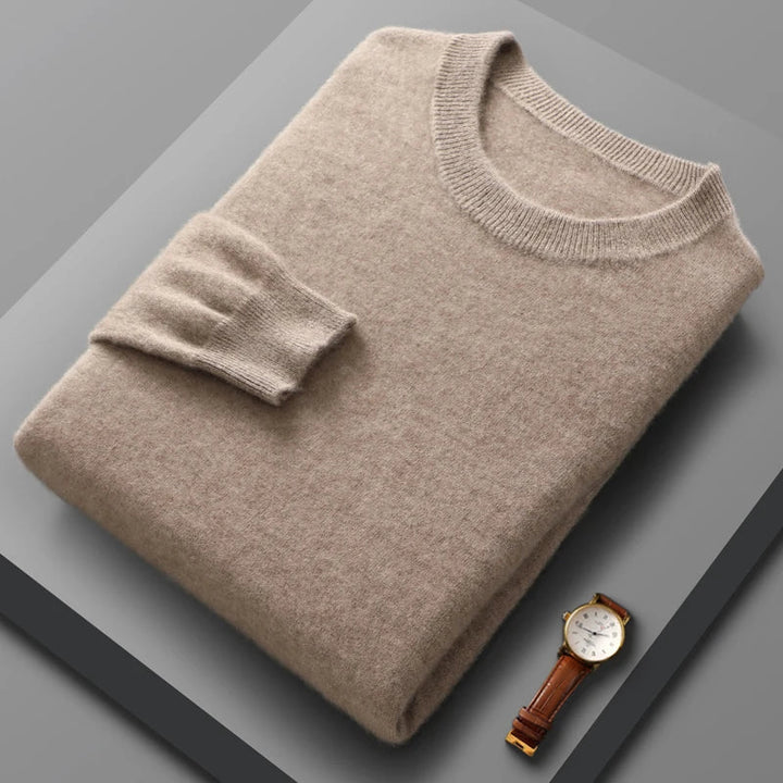 A neatly folded pure wool crewneck sweater with one sleeve draped over the front is placed on a gray surface; a classic wristwatch with a brown leather band lies next to it.