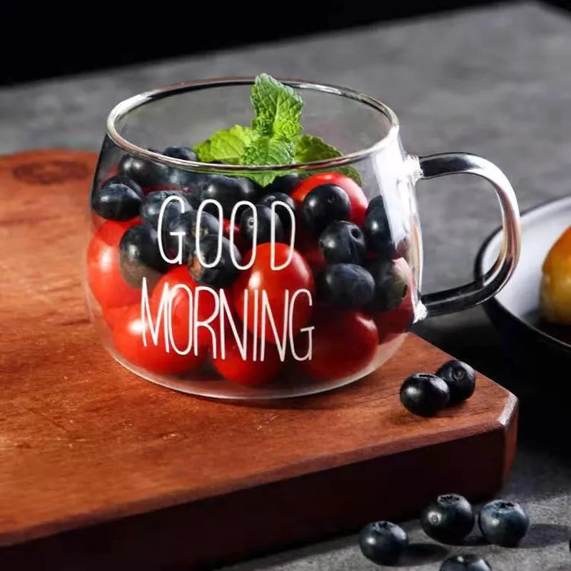 A clear glass mug with GOOD MORNING written on it, featuring an ergonomic handle, is filled with cherry tomatoes, blueberries, and a sprig of mint. It sits on a wooden surface with a few blueberries scattered nearby.
