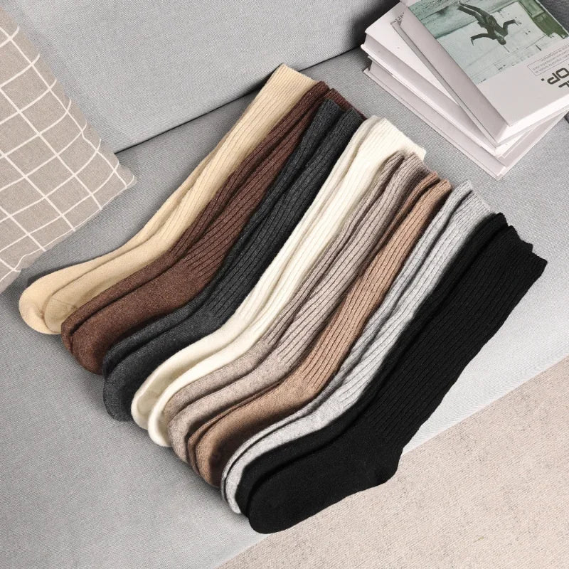 A row of ribbed, breathable socks in various neutral colors—cream, beige, brown, gray, and black—made from a soft Cashmere cotton blend, neatly arranged on a light gray couch next to a checkered pillow and books.