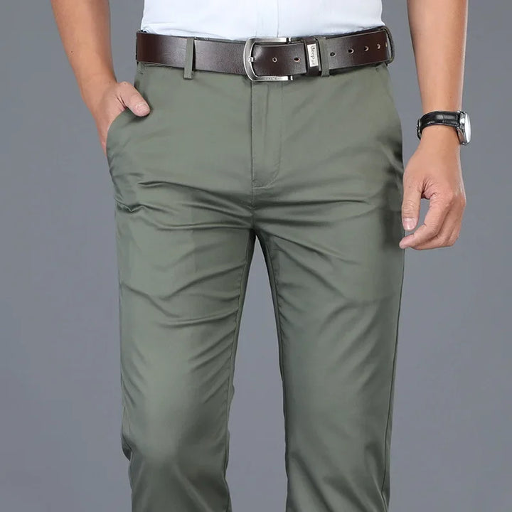 A person wearing olive green pants with a tailored fit, a brown belt, a white shirt, and a black wristwatch stands with one hand in their pocket against a plain gray background.
