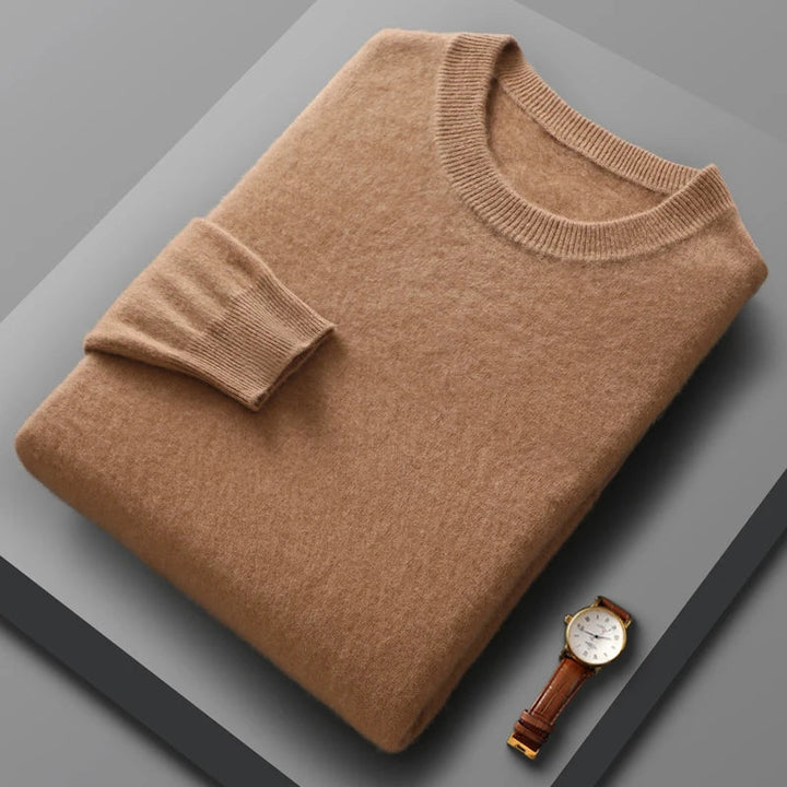A neatly folded light brown crewneck sweater with a ribbed collar and cuff, showcasing fine wool craftsmanship, rests on a gray surface. Beside it lies a brown leather strap wristwatch with a white face and gold case.