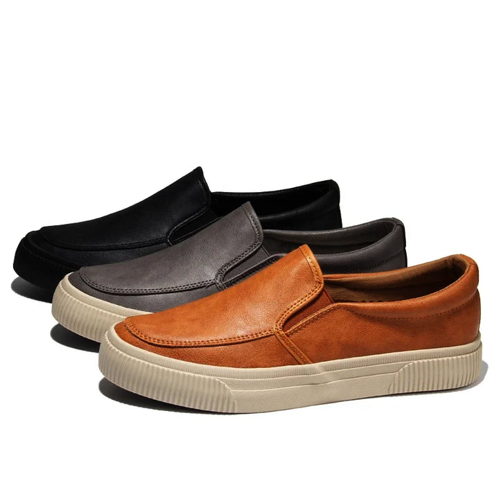 Three men’s slip-on sneakers are displayed side by side in black, gray, and brown. Each features a white hybrid rubber sole and a casual design with a comfortable leather insole, photographed against a plain white background.