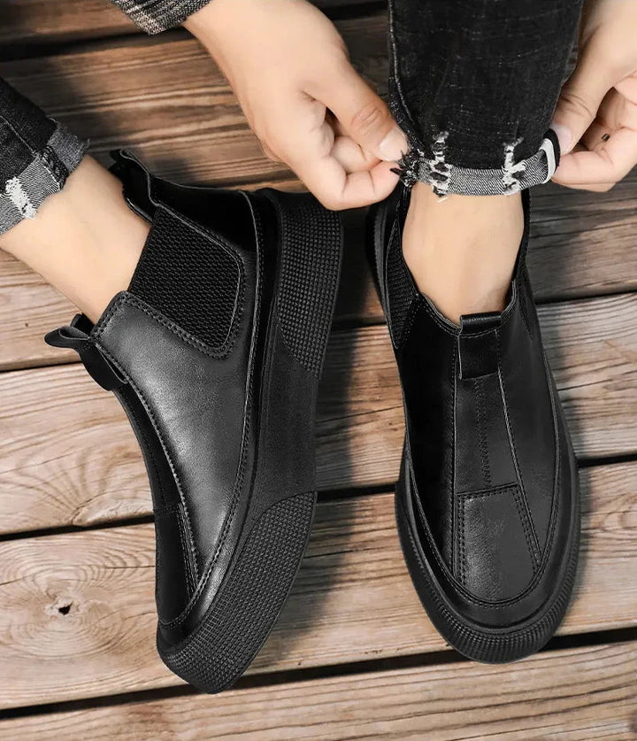 A person wearing black leather slip-on shoes with thick rubber soles and a cushioned footbed adjusts the cuff of their black jeans while standing on a wooden floor.