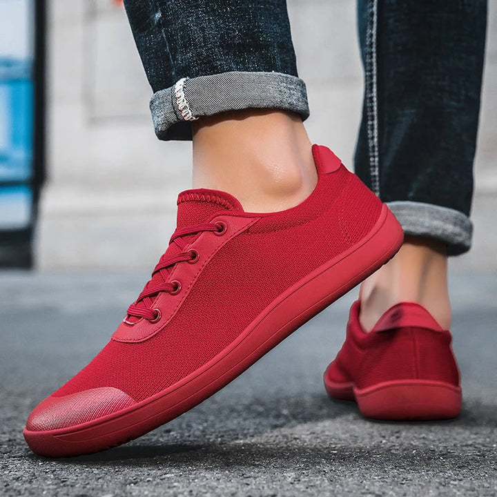 Person wearing rolled-up jeans and vibrant red men's canvas sneakers with a zero drop sole, walking on a paved urban street. The focus is on the shoes, highlighting their unique texture and striking color.