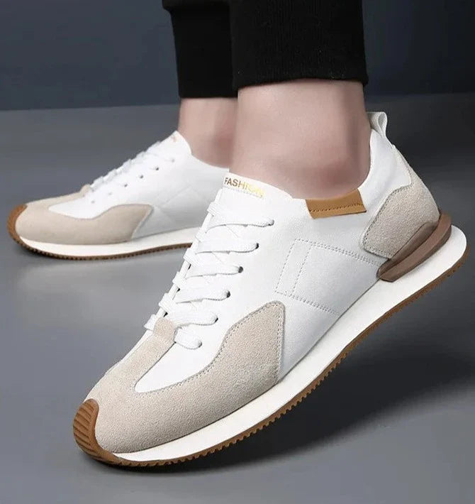 A person wearing stylish white and beige sneakers with brown accents and white laces, designed with top-grain leather for comfort, stands on a dark surface.