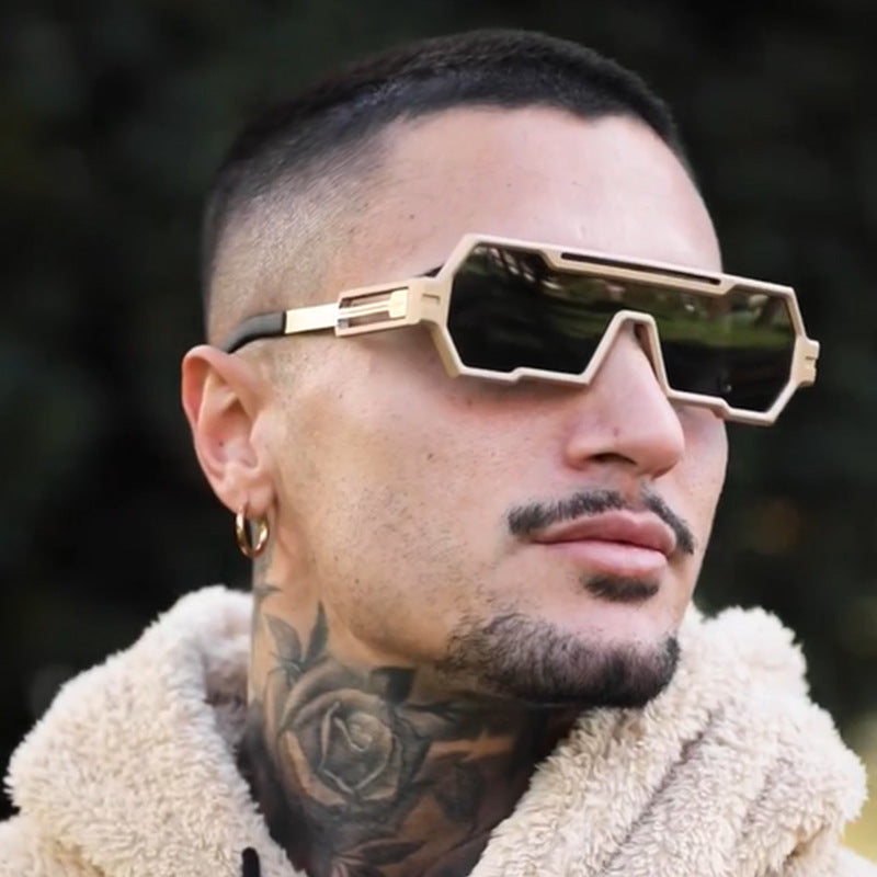 A person with short dark hair, a rose neck tattoo, a gold hoop earring, and a trimmed beard wears large square frames and a beige fuzzy jacket, looking off to the side against a blurred outdoor background.