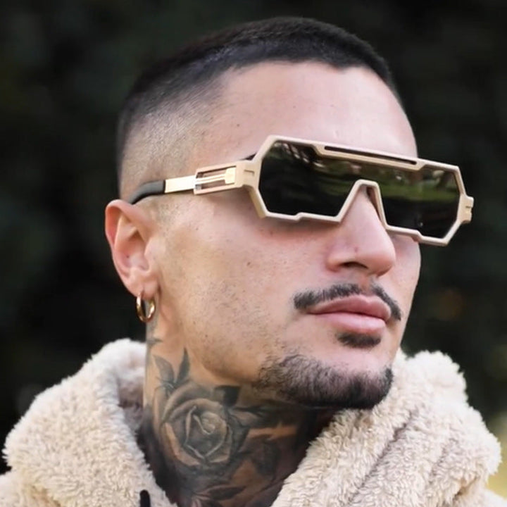 A person with short dark hair, a rose neck tattoo, a gold hoop earring, and a trimmed beard wears large square frames and a beige fuzzy jacket, looking off to the side against a blurred outdoor background.