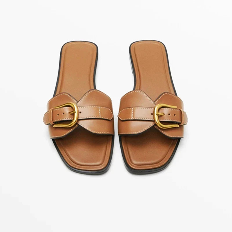 A pair of brown leather slides with large gold buckles and square toes, displayed on a white background.