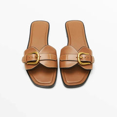 A pair of brown leather slides with large gold buckles and square toes, displayed on a white background.