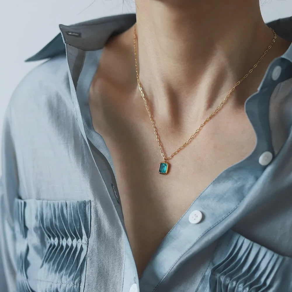 A person wearing a light blue, partially unbuttoned shirt with pleated pockets and an 18K gold plated chain necklace featuring a small rectangular green zirconia pendant.