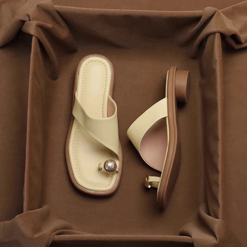 A pair of stylish beige and brown Square Toe Slides with a toe ring design, featuring a decorative stone on the toe strap, displayed on a brown fabric background.
