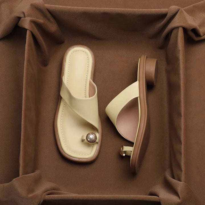 A pair of stylish beige and brown Square Toe Slides with a toe ring design, featuring a decorative stone on the toe strap, displayed on a brown fabric background.