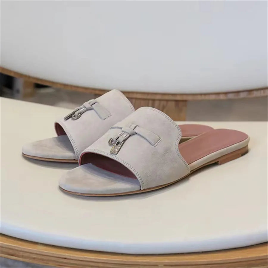 A pair of light beige suede finish slide sandals with open toes, flat wooden soles, cushioned insoles, and metallic buckle details on the upper straps, displayed on a cream-colored surface.