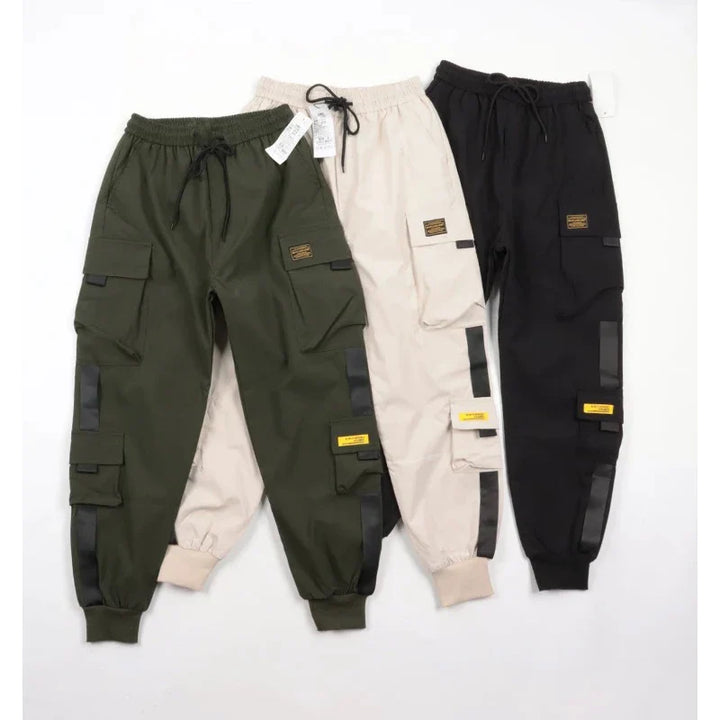 Three pairs of cargo pants in green, beige, and black arranged side by side. Each pair features an elastic waist, ribbed ankle cuffs, multiple pockets, black drawstrings, and decorative patches on the legs.
