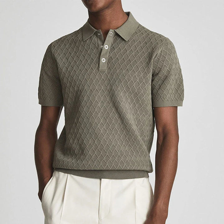 A person wearing a textured, olive green wrinkle-resistant classic polo shirt with a diamond pattern and cream-colored trousers, standing with hands in pockets against a plain background.