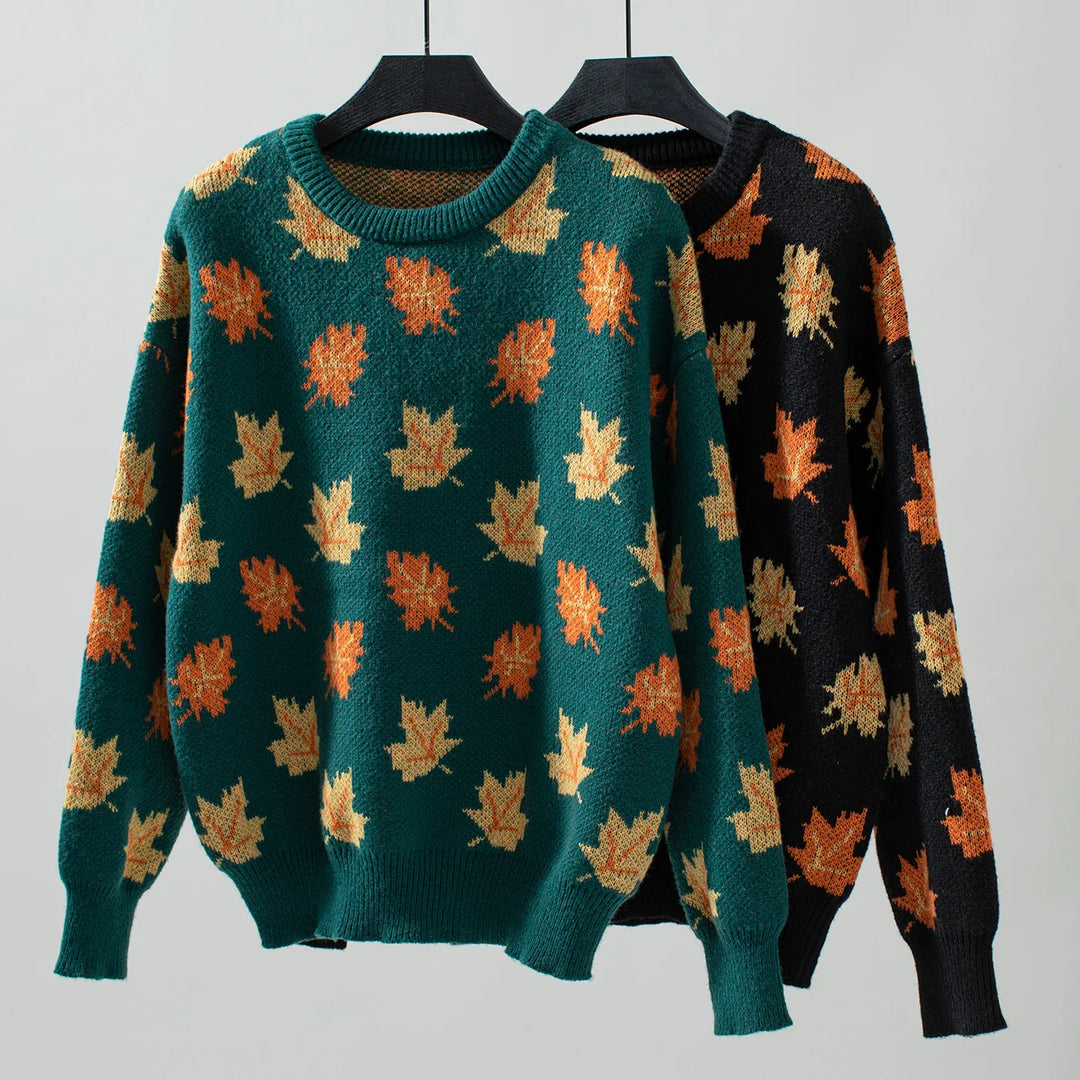 Two sweaters on hangers, one green and one black, both crafted from soft knit material and featuring an autumn maple leaf pattern in shades of orange and yellow, displayed against a plain light background.