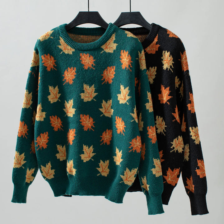 Two sweaters on hangers, one green and one black, both crafted from soft knit material and featuring an autumn maple leaf pattern in shades of orange and yellow, displayed against a plain light background.